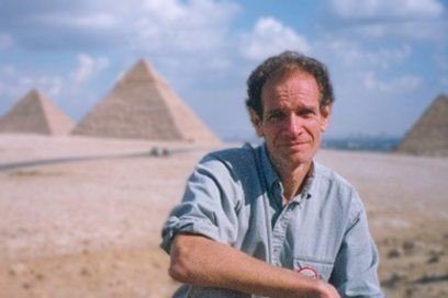 Dr. Bob Brier, crouching down in front of a trio of pyramids and blue sky