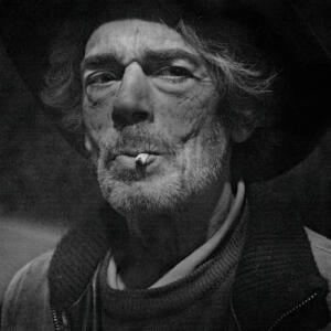 An older man with worn, grizzled features holds a cigarette tight between his pursed lips.