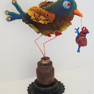 A blue and yellow bird made of various materials dangles an anatomic heart from its beak. At the start of its wire legs are 2-dimensional red heart shapes, and the ends of the legs stick into a tall, rusty geared part.