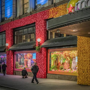 Several people in dark cold-weather clothing are on the sidewalk outside a store, the Macy's logo sticking out at the side. The outside wall of the bottom floor of the store is covered completely with yellow and red flowery material. The man in the foreground walks briskly with a hood on, and one man crouches at a display window where mannequins are dressed in nice dresses in a flowery scene.
