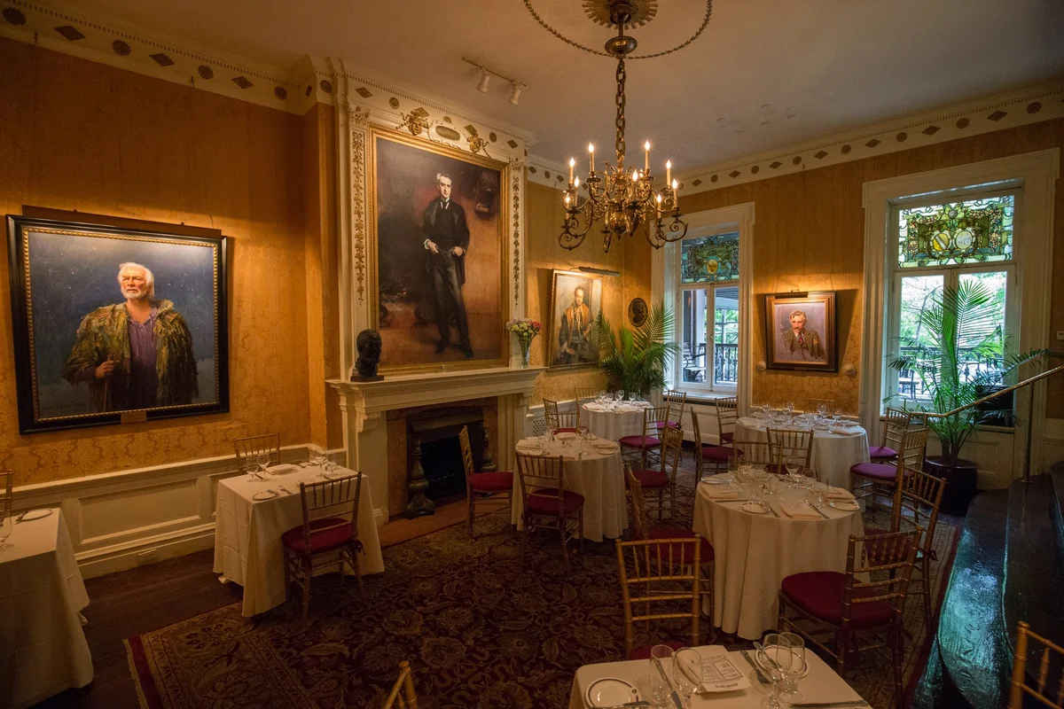 A dining hall of paintings, mustard yellow walls, and red seated chairs inside The Players Club.