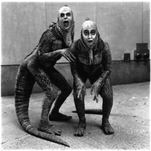 In black and white, two performers dressed in Godzilla-type costumes and pale facepaint pose playfully behind a building.