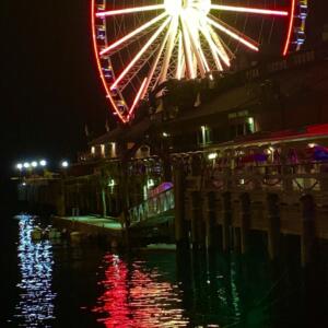 A ferris wheel near a pier shines brightly in the night, its neon reds and yellows reflecting in the water.