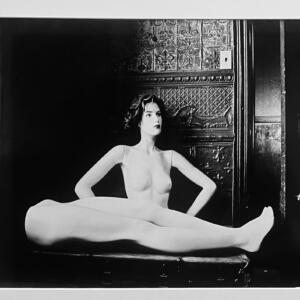 Black and white: the bottom half of a nude mannequin lays horizontally in front of her top half, who poses arms akimbo, looking up and away.