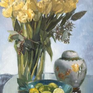 A large glass vase of yellow flowers, limes in a bowl,, and a round, ornate vase all on a circular, reflective platter.