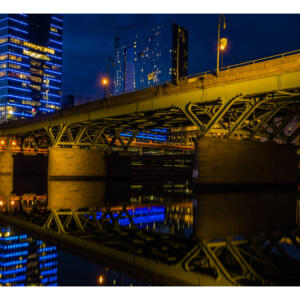 A city bridge over a body of water that reflects the brightly-lit skyscrapers ahead at night.