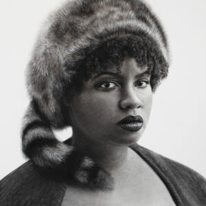 Black and white: a woman with dark lipstick wearing a Coonskin hat with a serious expression.