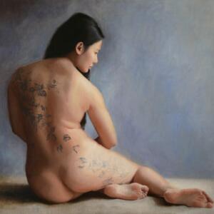 A nude woman with floral tattoos down her back and leg sits with her legs to her side. See see her from behind, her head turned to the side.