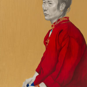 On a golden yellow background, an Asian man in grayscale sitting on a stool wears a bun in a red hairband, a small, gold hoop earring, a red shirt with a decorated collar, a red beaded bracelet, and blue pants.